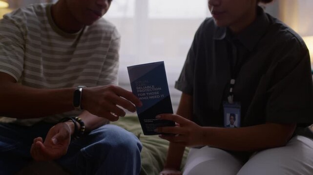 Midsection slowmo shot of African American teenage boy sitting on bed and talking to young female social worker in uniform giving him brochure with social assistance programs