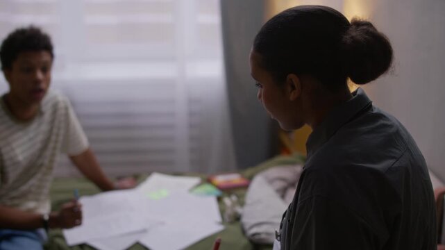 Tilt up shot of young Black female social worker with name badge taking notes during home visit while supervising teenage boy sitting on messy bed and doing school tests