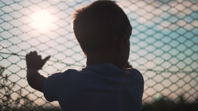 Boy holds fence while looking through chainlink barrier. Child stands watching sunlight behind fence outdoors. Silhouette of boy holding chainlink. Child looks through barrier at bright sunlight.