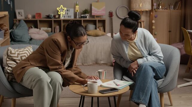 Medium shot of parent listening to female tutor who talking about childs grades and progress, they sitting in chair in girlish bedroom