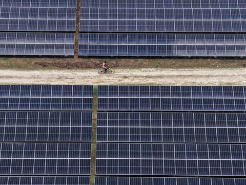 Aerial view of a lone bicyclist traversing a dusty path, framed by the geometric precision of solar panels stretching into the horizon, Gaibandha, Bangladesh.