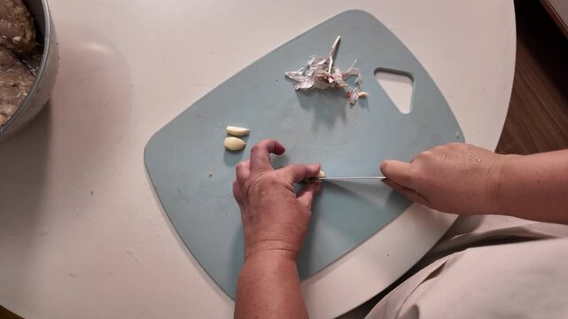 A woman's hands peel garlic with a knife on a cutting board