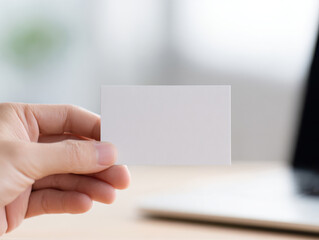 Obraz premium Close up of hand holding blank business card with blurred laptop and desk background for creative design or text