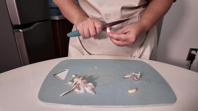 Woman in an apron peels a garlic clove over a cutting board.