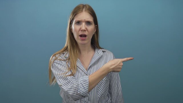 Blonde woman pointing finger to side with furrowed brow and tense posture in blue studio; frustration.
