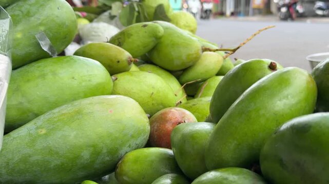 Fresh green mangoes fill a container prepared for sale, with urban street trade and vendor rows visible around the market space. Fruit distribution.