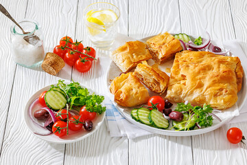 Bougatsa phyllo pastry with minced meat on white plate with red onion, olives and cherry tomatoes, cucumbers, Greek cuisine, horizontal view, not AI generated