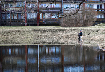Naklejka premium Senior Man Fishing Alone By Urban Pond With Apartment Buildings Reflection