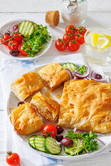 Greek meat bougatsa of crispy phyllo pastry with minced meat filling on plate with cucumbers, tomatoes, kalamata olives, red onion, parsley on white wooden table, vertical view, not AI generated