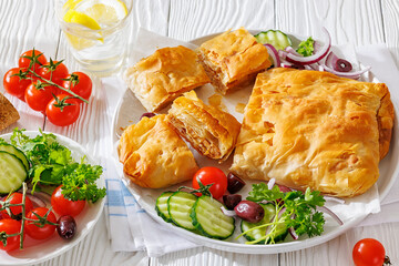 Bougatsa phyllo pastry with minced meat on white plate with red onion, olives and cherry tomatoes, cucumbers, Greek cuisine, horizontal view, close-up, not AI generated