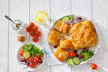 sliced Bougatsa phyllo pastry with minced meat on white plate with red onion, olives and cherry tomatoes, cucumbers, Greek cuisine, flat lay, free space, not AI generated