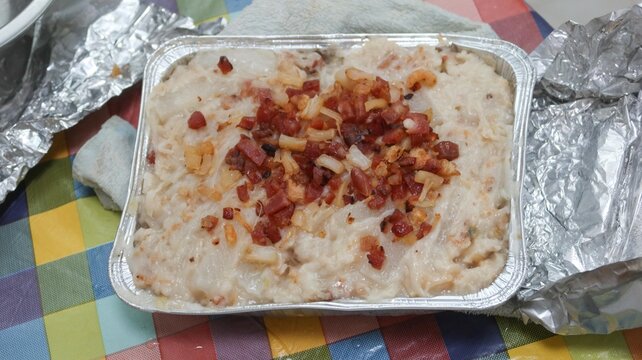 close up of Hong Kong traditional style homemade Chinese steamed turnip cake topped with Chinese sausage, dried shiitake mushrooms and dried shrimp in a Foil container on a white plate over colorful c