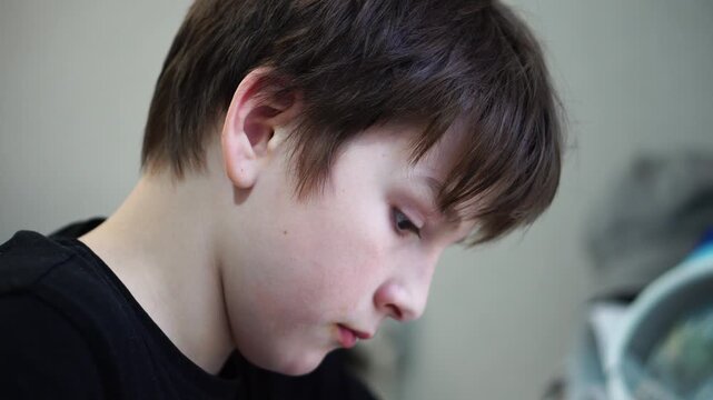 Close-up of a focused 12-year-old boy doing homework, writing in a notebook and looking at a computer screen.