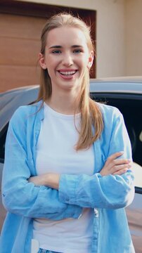Portrait of young woman standing near parked car in the parking looking at camera smiling. Girl charging electric car at charging station near home. Slow motion.