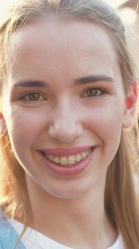 Close-up. Young beautiful girl smiles and looks at the camera against the background of pleasant sunlight at sunset. Portrait of attractive young adult stylish woman. Slow motion.