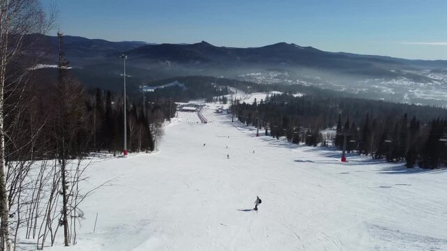 A drone view of a ski slope among tall trees, a ski piste at the Sheregesh resort. A snowboarder rides smoothly and softly on his board, evoking the atmosphere of freeriding and a love of the mountain