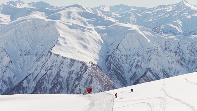 Aerial view Red snowcat drops riders on high alpine ridge, snowboarders prepare for freeride descent, vast Caucasus mountain range background, winter adventure tourism concept