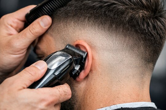 A close-up image of a man getting a haircut with an electric hair clipper near the ear, showing detailed grooming and hair trimming.