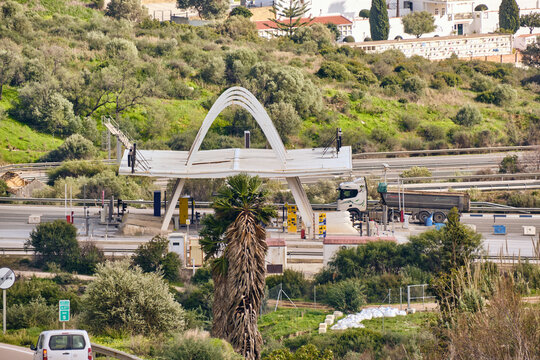 Manilva, Spain - 18 February 2026: View of the toll booth with its white arch standing in stark contrast to the green hills, trucks waiting, and the vibrant blue sky above.