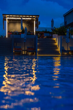A luxurious rooftop bar and pool area at night. The blue water of the pool reflects golden and lights from the structure above. A small bar pavilion with bright white LED outlines sits at the top