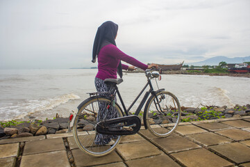 Fototapeta premium unrecognizable people, a woman wearing a hijab rides a vintage bicycle on the beach with mountains visible in the distance in the background.