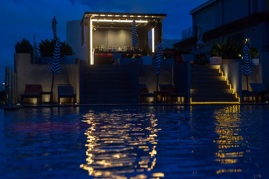 A luxurious rooftop bar and pool area at night. The blue water of the pool reflects golden and lights from the structure above. A small bar pavilion with bright white LED outlines sits at the top