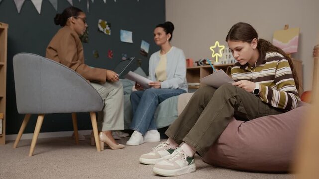 Caucasian teen girl sitting in bean bag chair and doing math test while her mother and female tutor communicating in background