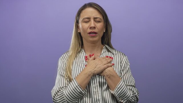 Woman clutch chest for heartattack in purple studio wearing striped blouse with red nails and pearl earrings; concern.