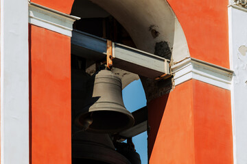 Architectural fragment of old bell tower featuring prominent metal bell set within vivid orange and white structure. Strong sunlight enhances textures of aged masonry and metal surfaces.
