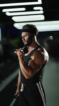 Focused bodybuilder performing intense dumbbell bicep curls in a modern indoor gym, displaying defined arm muscles, strength, and concentration under dramatic overhead lighting