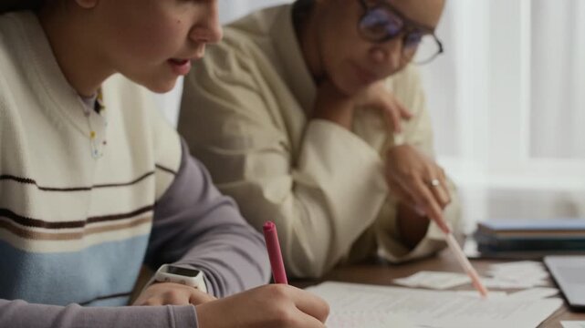 Close up of teen student writing on sheet of paper while her tutor guiding her and supporting