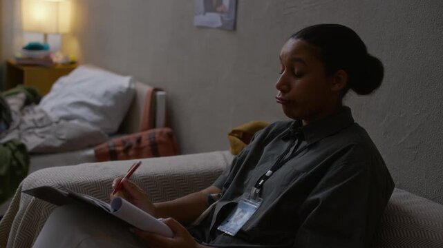 Slowmo handheld shot of young African American female social worker in uniform taking notes during home visit, supervising gen Z boy doing homework on messy bed