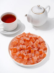 Orange Turkish delight cubes on plate with cup of tea and teapot on white background, sweet snack, tea time, studio still life