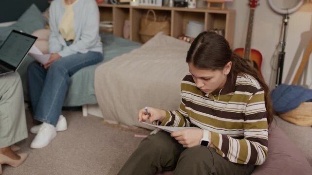 Math young tutor with laptop on laps having conversation with parent, discussing progress of teen girl who sitting in bean bag chair and doing test