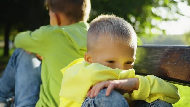 Younger brother looks sad with arms around knees while older brother sits turned away on bench. Concept of sibling rivalry and conflict