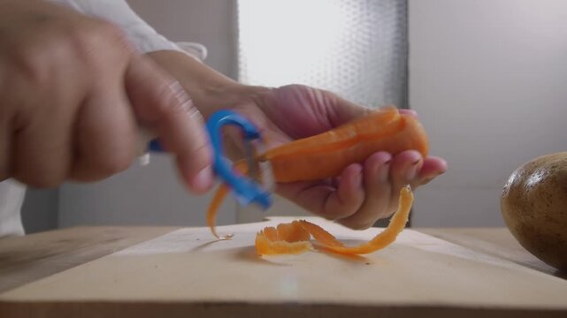 low angle view of woman's hands peel a fresh carrot with a vegetable peeler over a wooden table, preparing ingredients for cooking. Cooking preparation and healthy eating concept