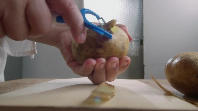 low angle view of woman's hands peel a potato with a vegetable peeler over a wooden table, preparing ingredients for cooking. Cooking preparation and healthy eating concept