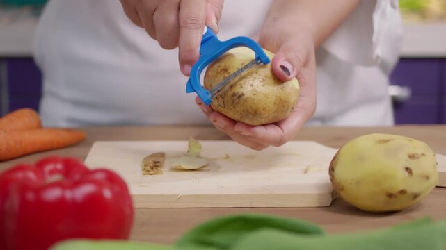 front view of woman hands peel a potato with a vegetable peeler over a wooden table, preparing ingredients for cooking. Cooking preparation and healthy eating concept