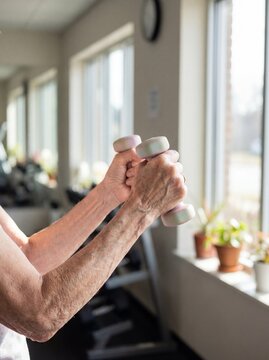 Sarcopenia (Muscle Loss): Professional photography, close-up of thin, frail elderly arms lifting a very small dumbbell, visible effort, bright morning gym setting.