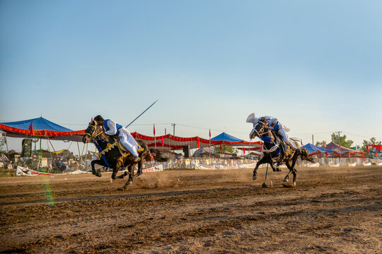 Punjab, Pakistan - 24 April 2024: View of two riders on horseback racing across the dusty ground, lances poised under a clear sky, amidst the vibrant colors of tents and flags.