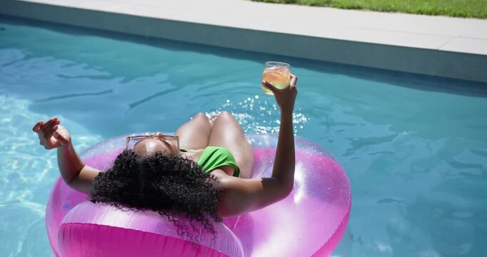 Woman in bikini lying on pink float in home pool holding drink friend reaching shading eyes