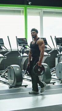 Muscular athlete performing a loaded carry with weight plates through a modern gym, walking past rows of cardio machines by windows, showing strength, grip, and conditioning