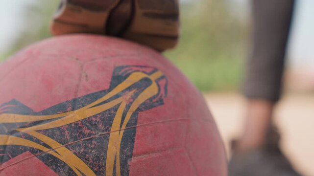 Black player shoe pressing worn ball emblem, shallow depth of field with blurred legs, textured scuff marks and logo detail, quiet preplay ritual, warm light, emotional grassroots focus.