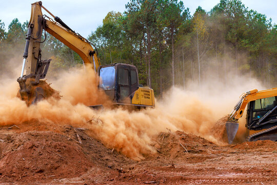 Construction skid steer equipment, excavator digs into ground, kicking up dust at site preparation land development.