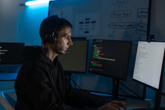 Male programmer and developer focused on computer screen at night, coding digital systems and ensuring cyber security. Freelancer with headset working overtime under blue light in office.