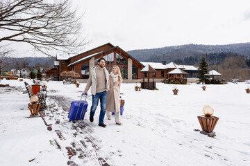 Happy couple arriving at a winter resort, pulling suitcases for a snowy vacation