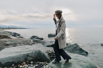 Woman in a grey coat and hat walks on rocks by the sea during cloudy day. Calm coastal landscape with pebbles and water in the background. Concept for autumn or outdoor lifestyle.