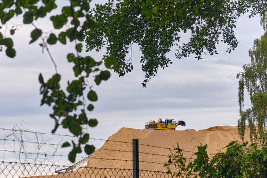 Nymolla, Sweden - 21 September 2025: View of a colossal mound of woodchips topped by a yellow bulldozer, framed by the delicate green foliage and a wire fence.