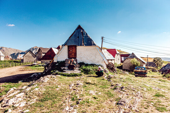 Lukomir, Bosnia and Herzegovina - 28 August 2015: View of an aged woman sitting in front of her traditional stone and wood house with a distinctive pyramidal roof, under a clear blue sky.