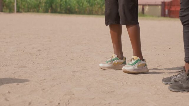 Two players trade passes on dirt pitch, one in black pants, closeup on feet and pink ball, dust puff with each touch, playful teamwork and footwork, grassroots match energy under warm summer light